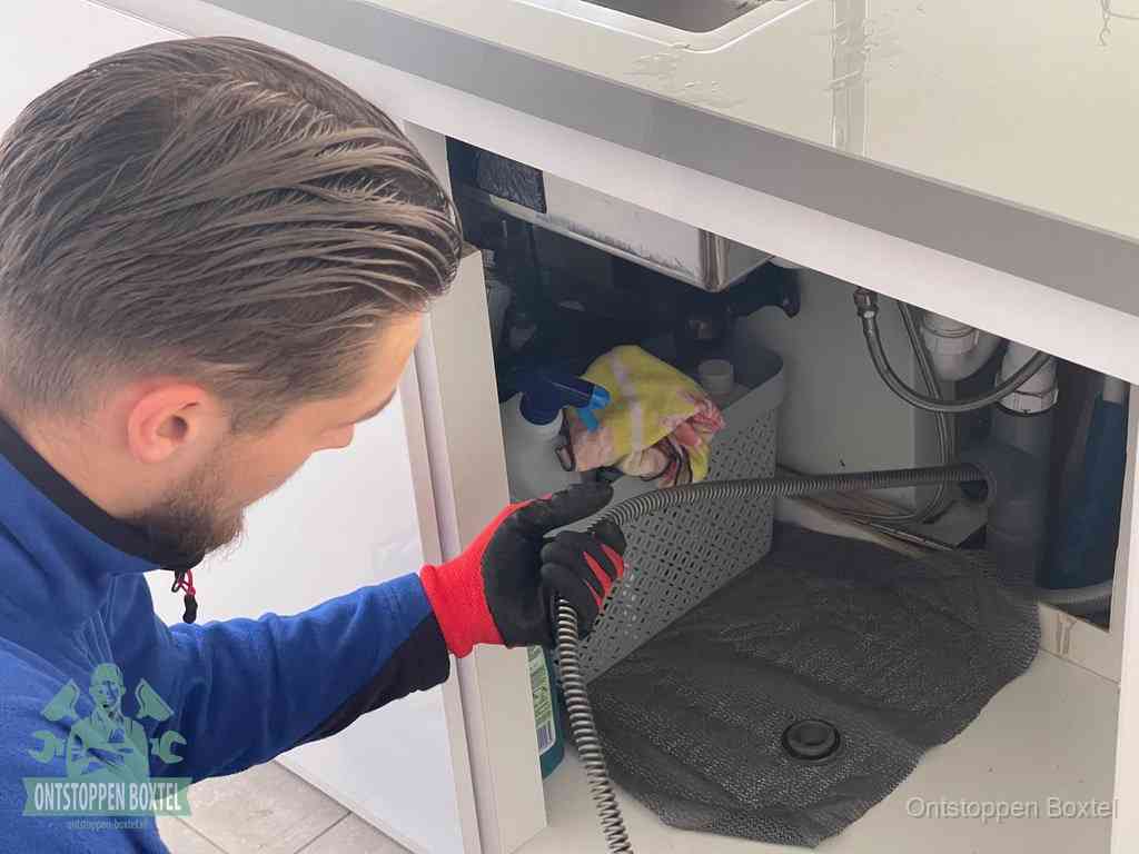 Een monteur voert een flexibele ontstoppingsveer in onder een keukenkastje om een verstopping voorbij de sifon te bereiken in Boxtel