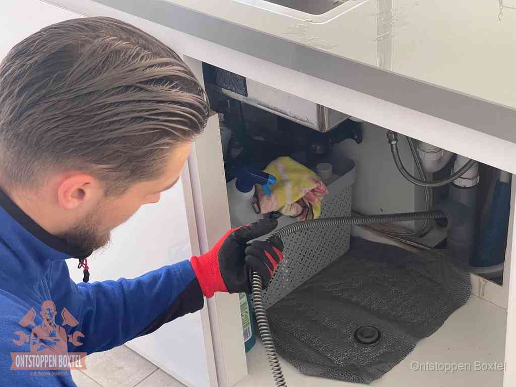 Een monteur voert een flexibele ontstoppingsveer in onder het keukenkastje, langs de sifon, om de gootsteen te ontstoppen in Boxtel