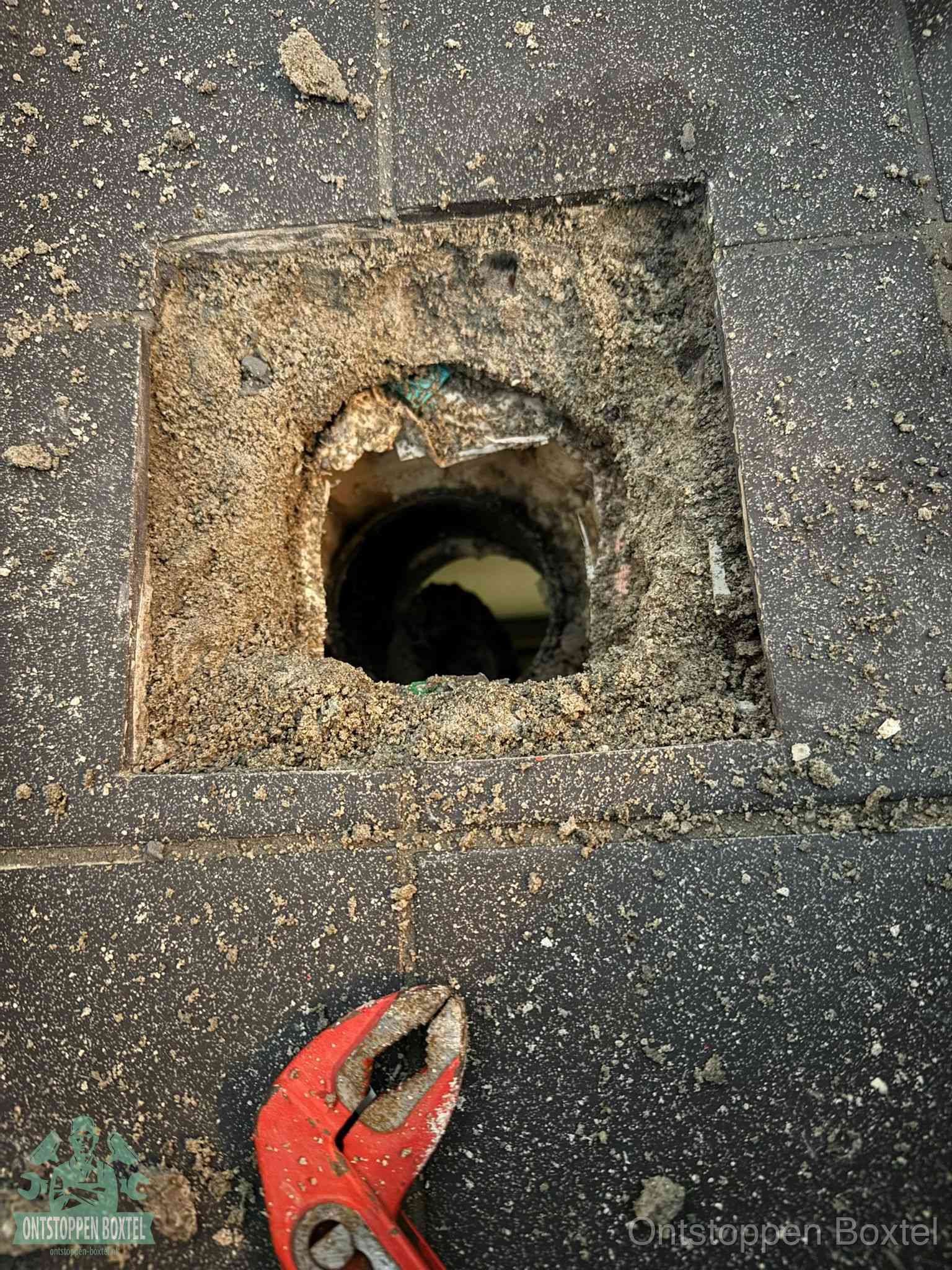 Een geopend rioolputje in een betontegelvloer met losgemaakt rooster en een zichtbare ronde afvoerbuis, het achterhalen waar de afvoerverstopping zit in Boxtel