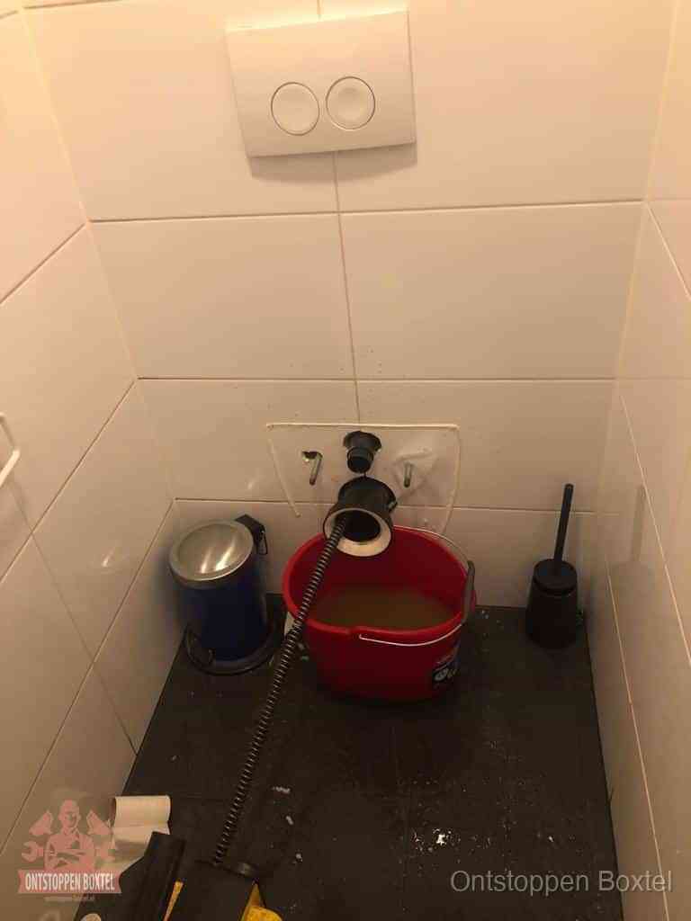 Een ontstopte wand-wc met verwijderde toiletpot waar een ontstoppingsveer in de wanduitgang verdwijnt, het achterhalen waar de verstopping zit in Boxtel