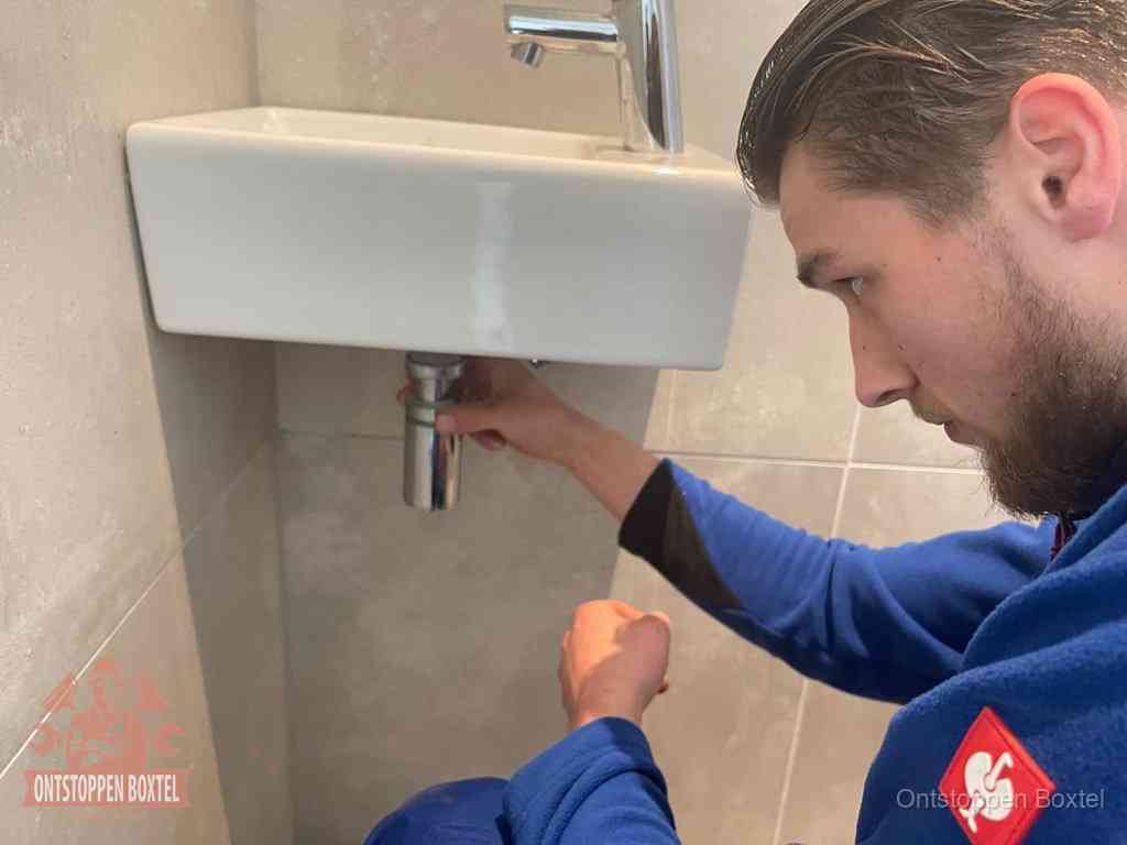 Een monteur draait met de hand de zwanenhalssifon onder een fonteinwasbak in de toiletruimte los om de bron van rioollucht in Boxtel te vinden