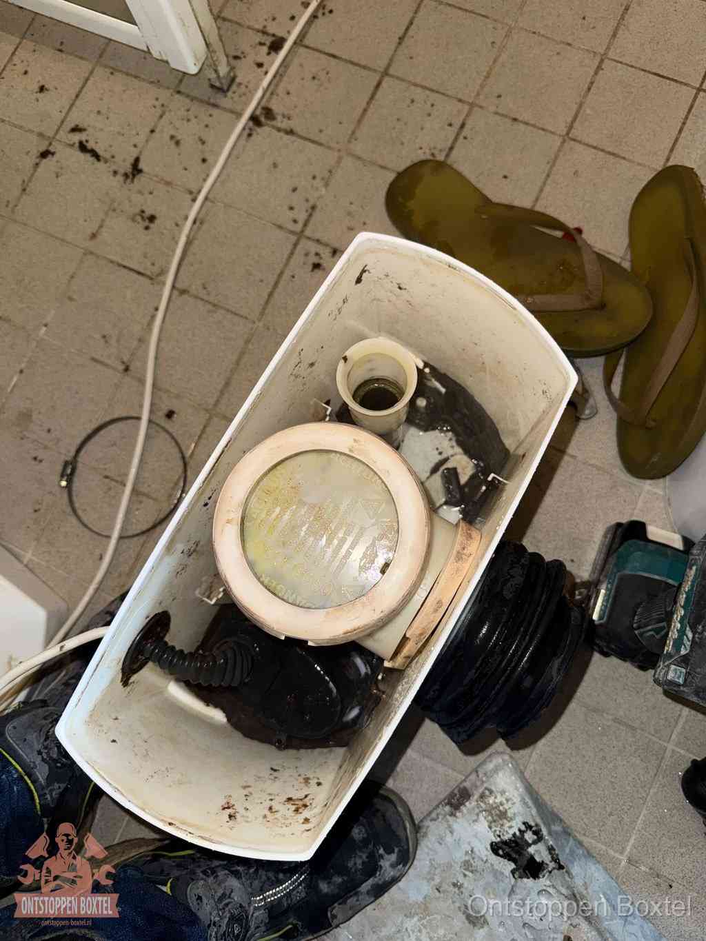 Een gedemonteerde toiletpot op de vloer met ontstoppingsveer en gereedschap ernaast, het verwijderen van een vastzittend voorwerp uit de wc in Boxtel