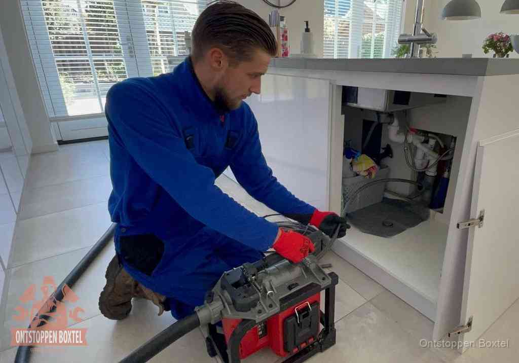 Een monteur voert een professionele elektrische ontstoppingsmachine in de afvoer onder de spoelbak tijdens het gootsteen ontstoppen in Boxtel